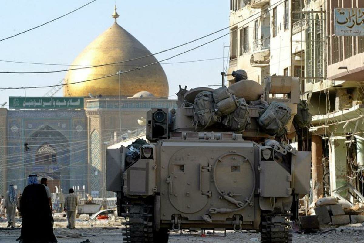 Rear view of a tan U.S. Army Bradley armored vehicle driving down a debris-filled street in Iraq, with civilians walking nearby and the golden dome of the Imam Ali Shrine prominent in the background.
