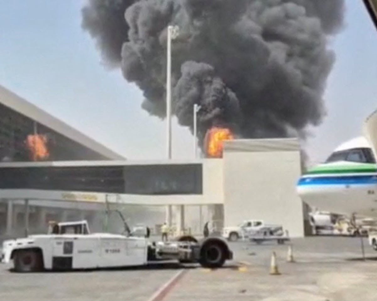 A large plume of thick black smoke rises from a major fire at an airport terminal structure next to the tarmac. A white airport tug vehicle sits parked in the foreground, and the nose of a commercial airplane with green and blue stripes is visible on the right