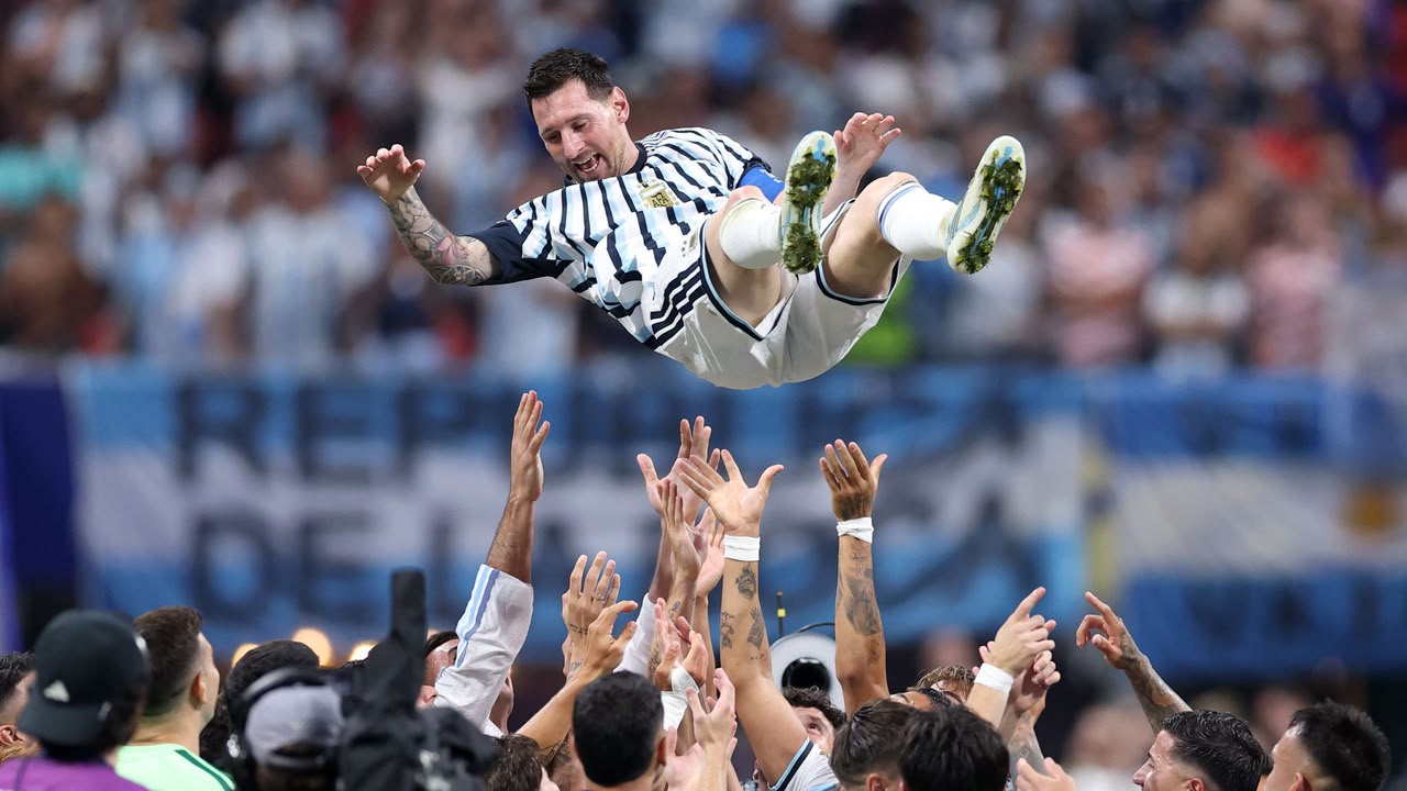 Argentina national football team players toss captain Lionel Messi into the air inside a crowded stadium to celebrate winning the 2021 Copa América final. Messi is airborne, horizontal, and smiling, while a crowd of teammates raise their arms underneath him against a backdrop of blurry spectators.