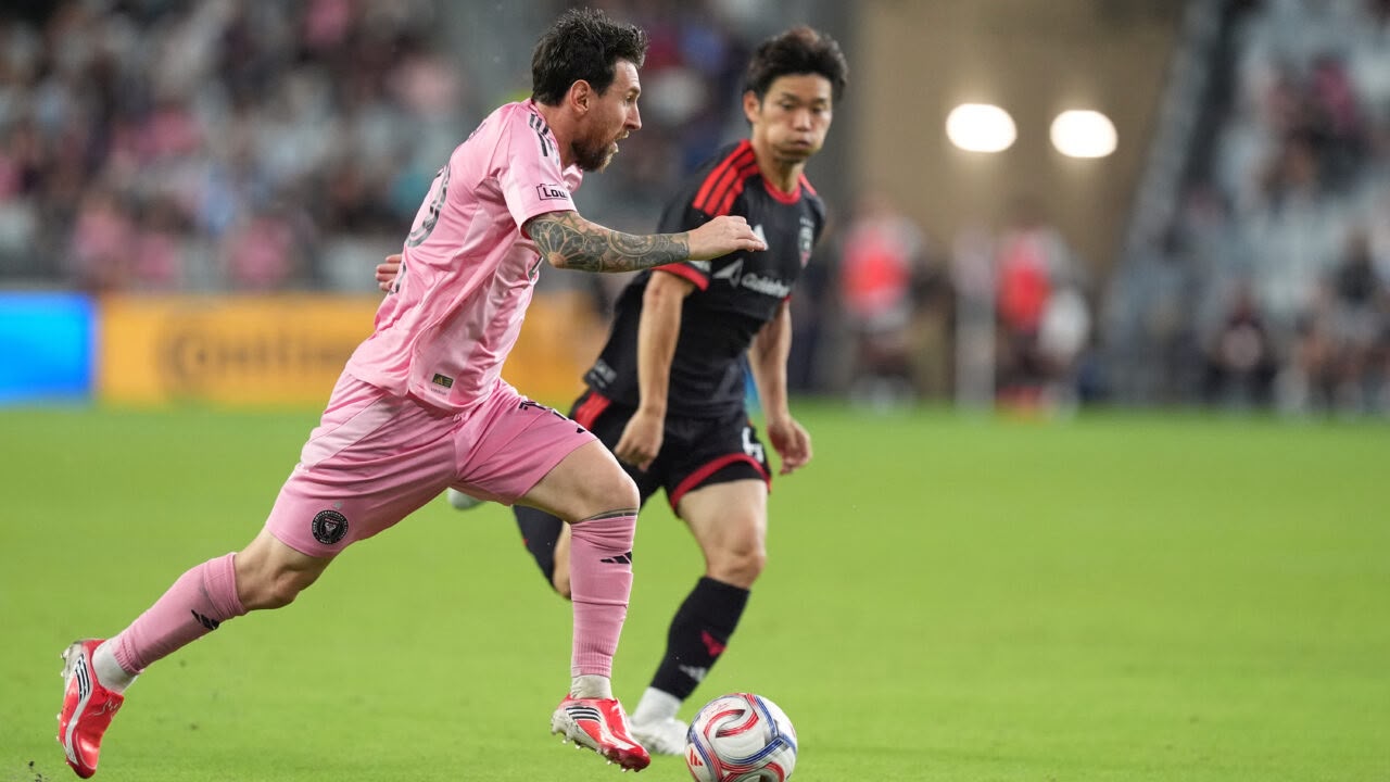 An action shot of Lionel Messi, wearing Inter Miami CF's signature all-pink uniform, dynamically controlling a soccer ball while running down the field as a opposing player in a black and red uniform trails closely behind.